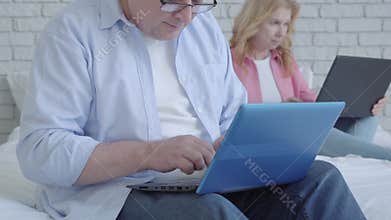 Smiling mid-adult man using social media on laptop and smiling with blurred senior woman surfing Internet at the
