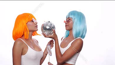 Two young smiling Asian women in bright color wigs posing with photo booth props and small disco ball over white