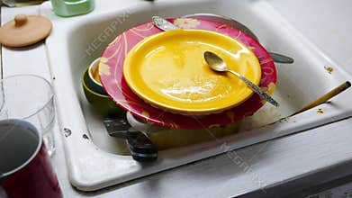 Stack of dirty plates, cups and spoons in a sink.