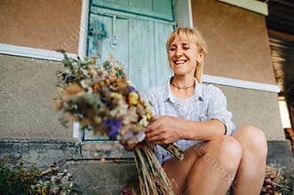 Portrait of a happy female florist sitting at the cottage in dried flowers and making a beautiful bouquet. Making bouquets of