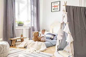 Cute little boy sitting in comfortable bed playing with teddy bear, real photo of natural playroom interior