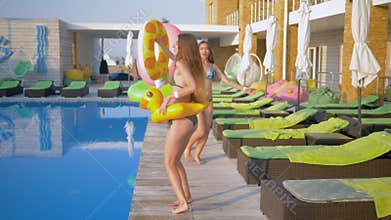 Happy attractive girlfriends into bathing suit with Inflatable rings walk near pool during summer holiday at resort