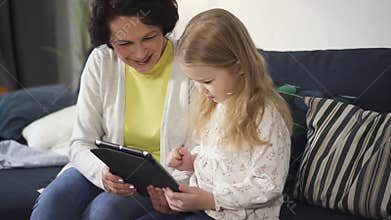 Mother or grandmother is sitting on sofa and showing to little girl digital technology