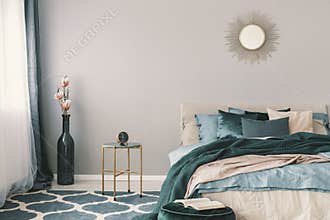 Flowers in stylish bottle like vase next to trendy nightstand with clock in beautiful bedroom interior with beige and emerald
