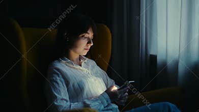 Young beautiful woman in casual clothing uses a phone in a dark room.