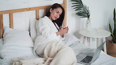 Woman in white bathrobe, lies on bed, drinks tea and watches movie on laptop