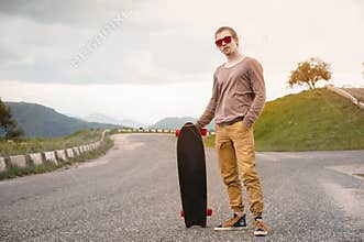 A stylish young man standing along a winding mountain road with a skate or longboard in his hands the evening after