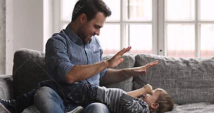 Caring young dad tickling child son laughing playing on sofa