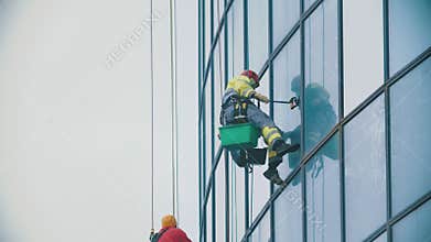 A man worker in yellow work clothes hanging on ropes and wipes the exterior windows of a business skyscraper - holding
