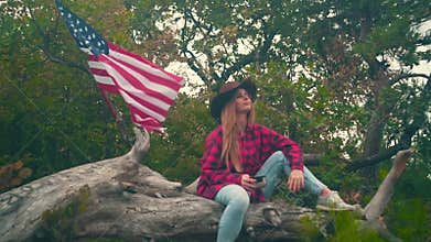 A girl in a cowboy hat in a plaid shirt sits on a log and drinks a tea. In the background, an American flag flutters.