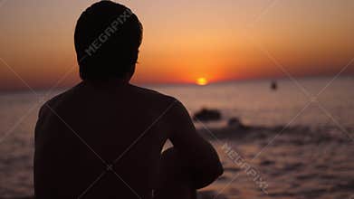 Silhouette of a man sitting alone on the beach looks sunset. Lonely, thinking person.
