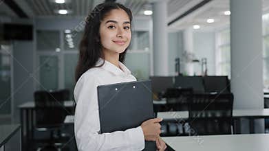 Young confident Indian businesswoman model posing indoors smiling female manager office worker trainee stands holding