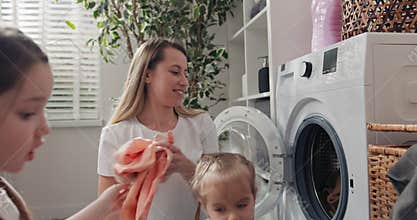 Taking clothes out of the wicker basket and putting them in the washing machine drum. Mother and children tidy up the
