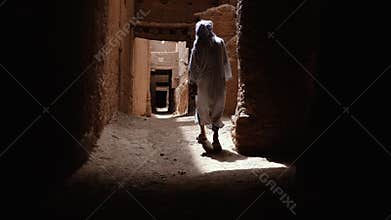 A traditional dressed Moroccan bedouin man wearing a white gandoura and a turban walks inside a Kasbah in Tamnougalt, Morocco.