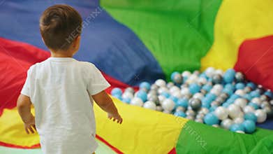 little Caucasian boy playing with a colorful piece of material and many balls on it, full shot kindergarten