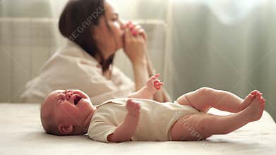 Tired infant girl cries on bed against depressed mother