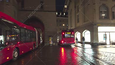 The center of Bern at night with buses, trams, statues and cathedrals with locals and tourists walking by
