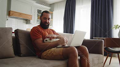 Adult man works with one hand on a laptop and also holds his little cute dog. An freelancer guy work from home online