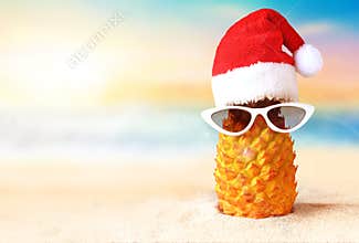 Pineapple in red Santa Claus hat in sunglasses on a sand on the tropical beach near ocean, summer Christmas concept