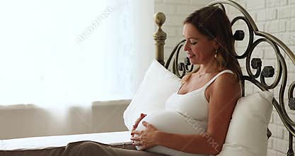 Peaceful expectant mother stroking her belly resting seated on bed