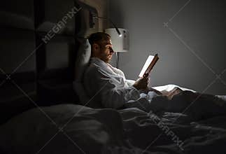 Midle-aged Caucasian man relaxing in bed reading bestseller novel paper book with bedside lamp turned on. Evening relaxation,