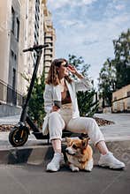 Young businesswoman in white suit sitting on electric scooter and using phone during walking with Welsh Corgi Pembroke dog in city