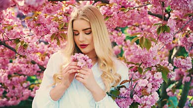 Happy blonde hair fashion model woman holding in hand pink sakura flowers, tree park spring nature garden sun light