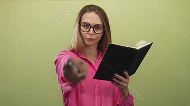 Young woman in glasses reading a book and pointing at the camera with an amused expression against a yellow background wall