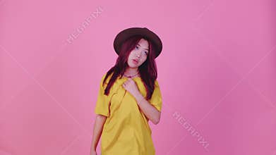 A young Asian woman with vibrant red hair, wearing a hat and a yellow t-shirt, stands against a pink background looking