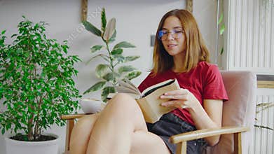 A young, beautiful girl wearing glasses is sitting in an old armchair in a cozy cafe, deeply engrossed in a book. The