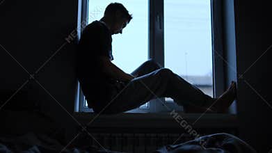 Silhouette of a man sitting on a windowsill, holding his head, showing sadness and emotional struggle in a dimly lit
