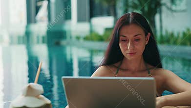 Young woman enjoys working holiday by hotel swimming pool using laptop at tropical resort.