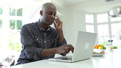 Man Using Laptop And Talking On Phone In Kitchen At Home