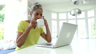Woman Using Laptop And Talking On Phone In Kitchen At Home