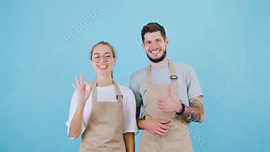 A man and a woman in aprons stand against a blue background, looking into the camera and giving a thumbs-up. The man has