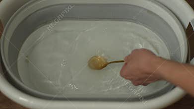 Close-up hands of unrecognizable man pouring washing powder into spoon over basin, adding to water and stirring