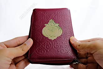 Hands holding a maroon Quran with gold emblem on white.