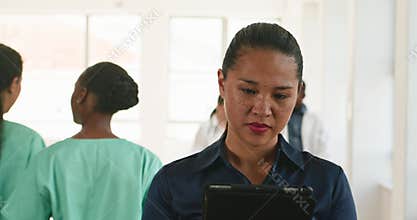 Dedicated Doctor Reviews Patient Information on Tablet in Busy Hospital Hallway with Medical Staff in Background