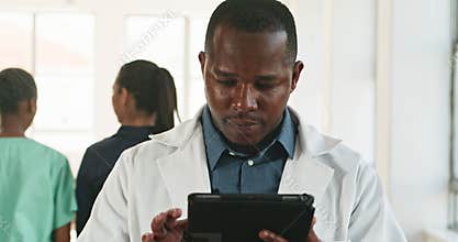 Male Doctor Reviews Patient Information on Tablet in Busy Hospital Setting with Colleagues in Background