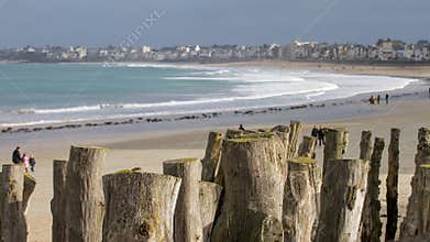 Visitors walk along the beach in Normandy with wooden posts marking the shore and waves gently crashing on a sunny day