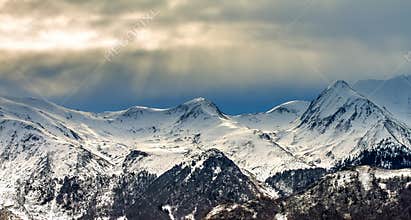 Snow-covered Pyrenees mountains under cloudy sky with sunlight breaking through in the evening