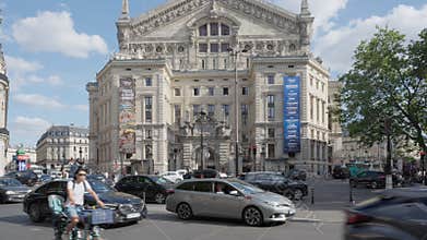 Vehicles and scooters drive through a busy intersection by Palais Opera Garnier