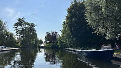 Quiet canal scene with moored boats, lush riverside trees, and people relaxing