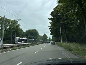Cars driving along tree lined urban road beside tram stop and rail