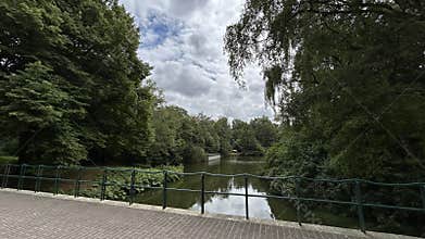 Quiet park lake framed by dense green trees, brick walkway, metal railing
