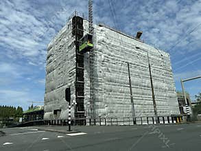Large wrapped building under renovation beside wide city road, with scaffolding