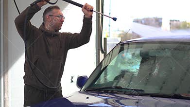 A middle aged man with eyeglasses washes a blue car at a self-service car wash.