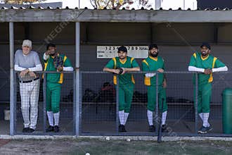 Diverse male baseball team and coach standing on dugout rail in green uniforms holding gloves, bat