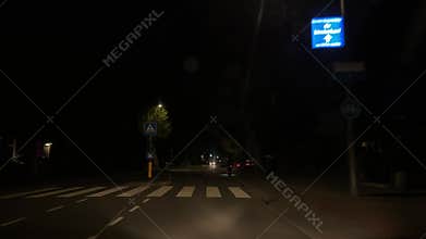 Empty night street with zebra crossing, headlights, parked cars, and roadside