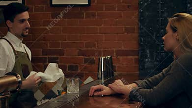 Smiling blonde woman listening to bartender, while he talking and rubbing glasses in bar